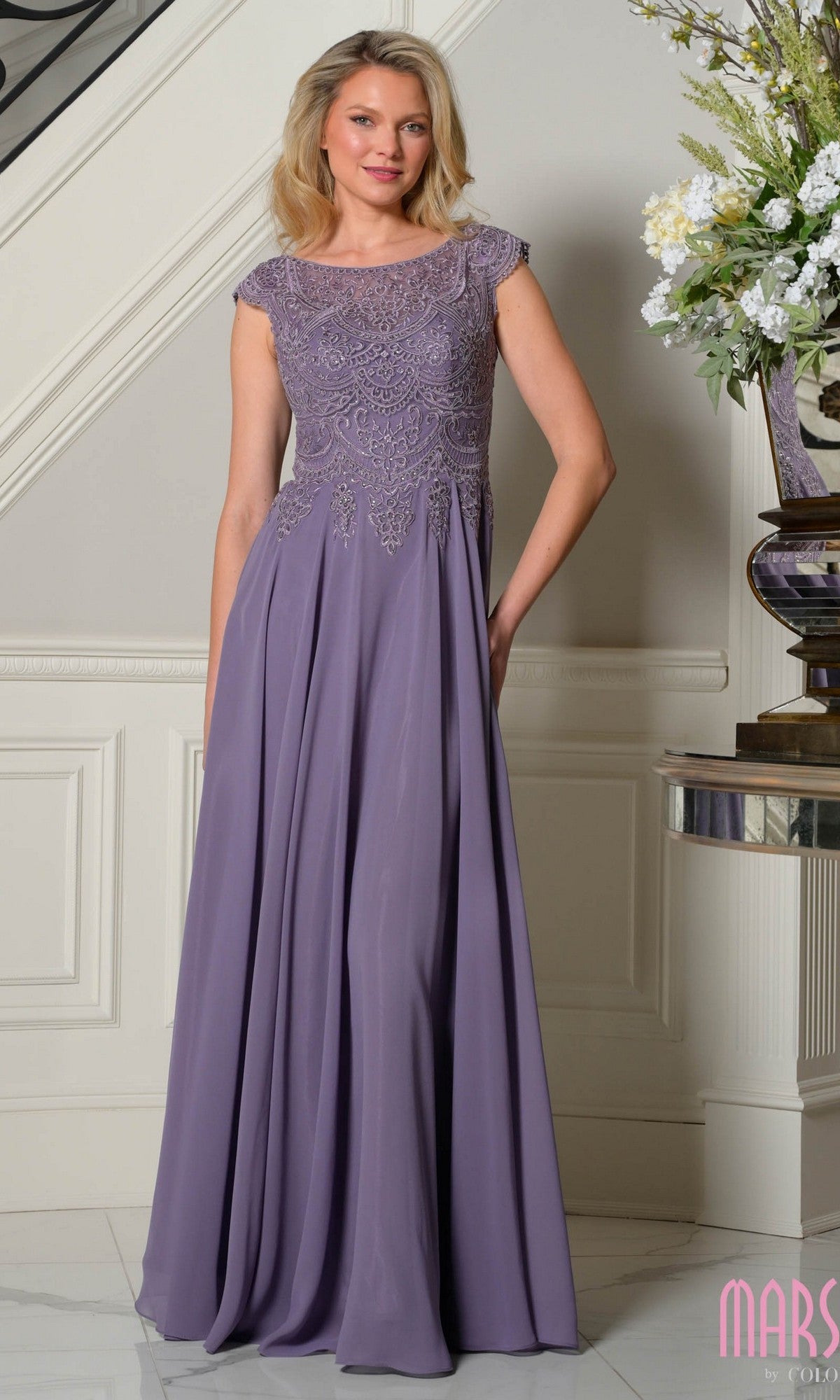 A woman stands indoors by a floral arrangement, wearing the Marsoni M238 long formal dress in wisteria with lace detailing on the bodice.